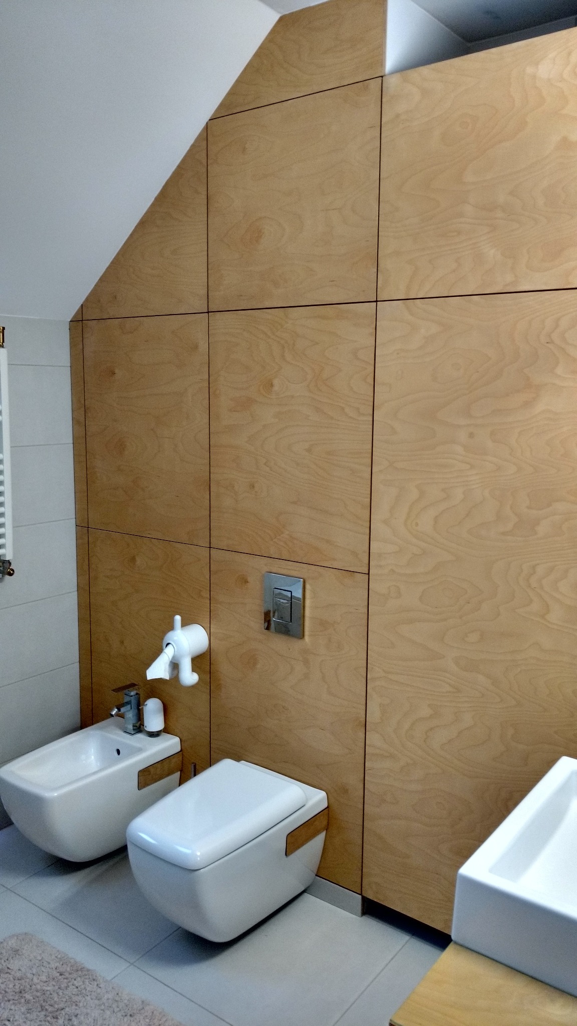 Nowoczesna łazienka z drewnianą zabudową ściany pod skosem, wisząca toaleta i bidet w minimalistycznym stylu, chromowany przycisk spłuczki.