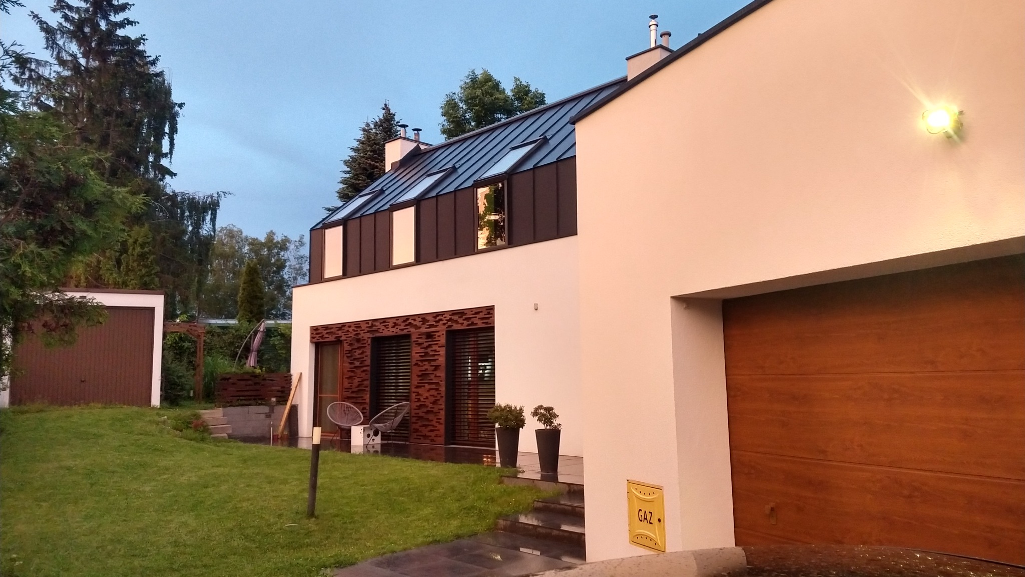 Nowoczesny dom jednorodzinny z elementami dekoracyjnymi z cegły, oświetleniem zewnętrznym i garażem z drewnianą bramą, otoczony trawnikiem i zielenią.