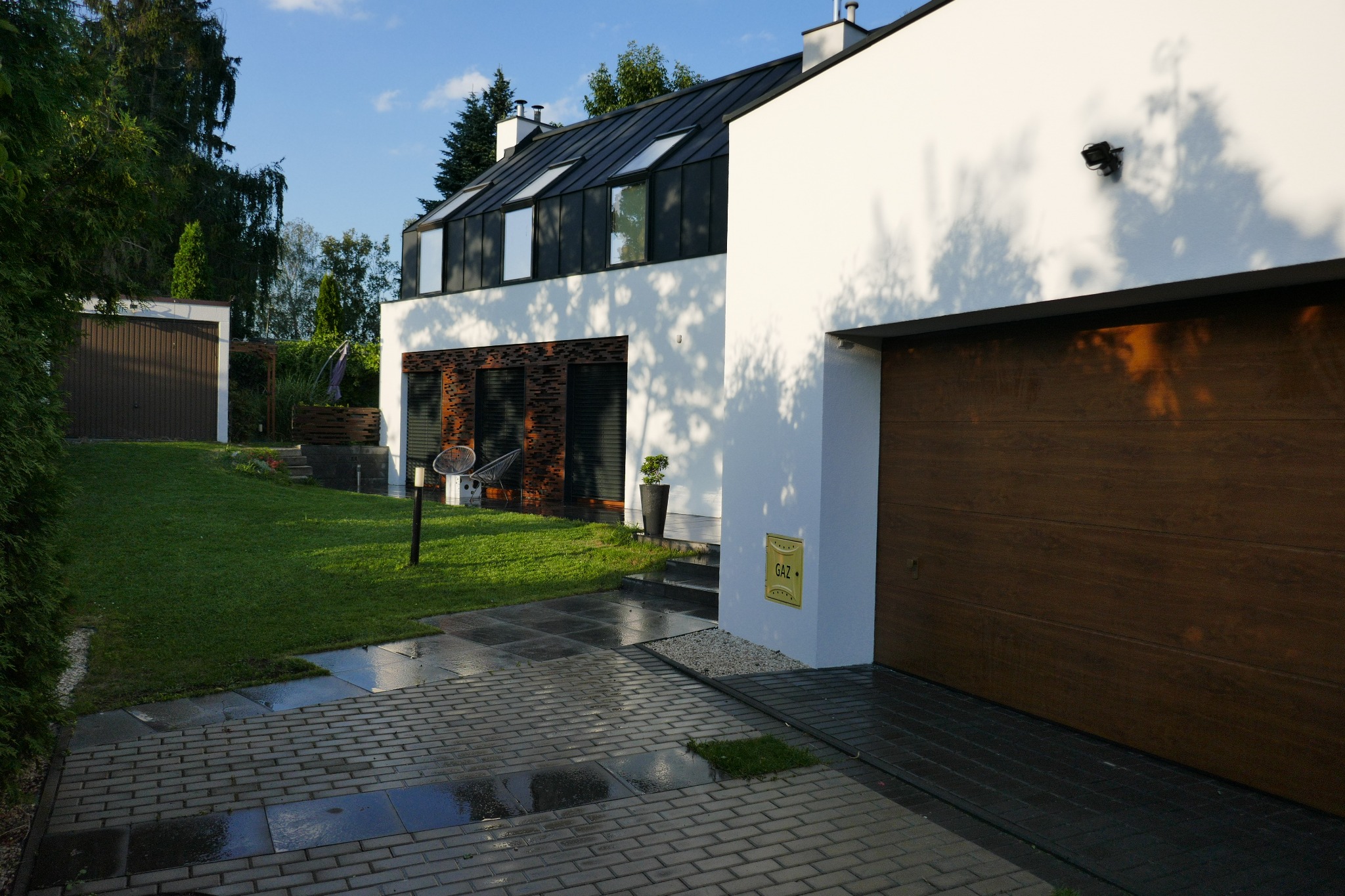 Nowoczesny dom jednorodzinny z garażem, biała elewacja, czarna dachówka z oknami dachowymi, ozdobna elewacja z cegły, zadbany trawnik, brama garażowa z drewna, kostka brukowa.