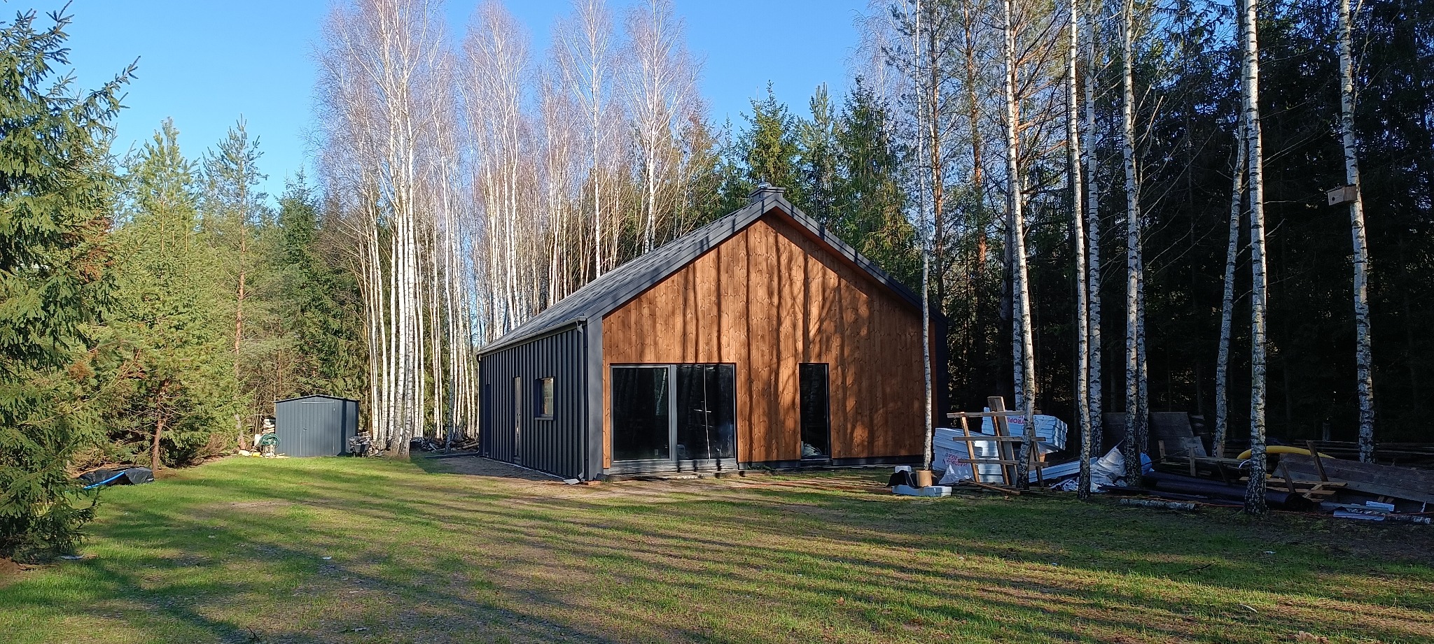 Nowoczesny dom jednorodzinny z drewnianą elewacją i ciemnymi oknami, otoczony lasem brzozowym, w tle widoczny blaszak i materiały budowlane.