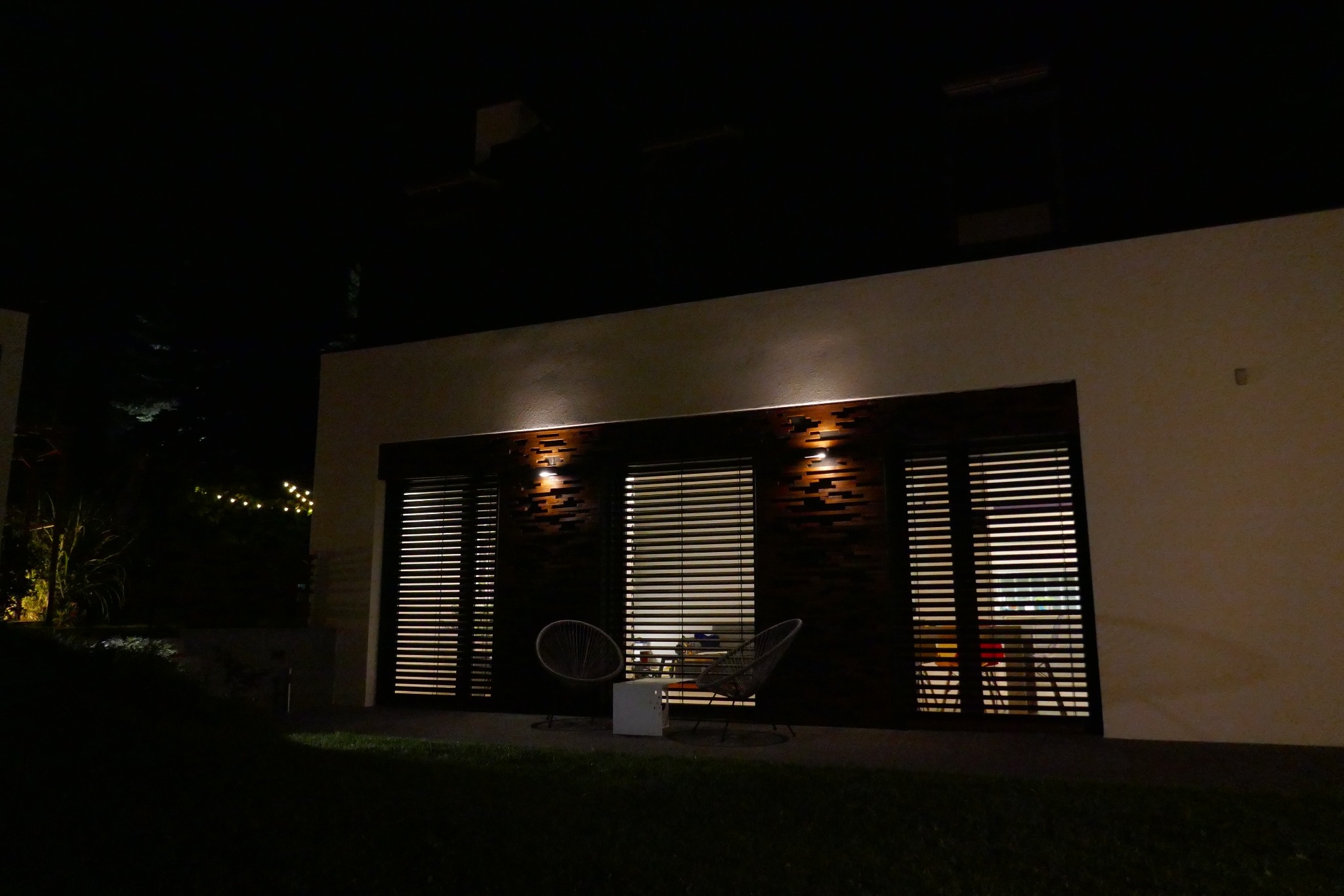 Nowoczesny budynek z drewnianą elewacją, oświetlony dwoma lampami, z widocznymi roletami w oknach i dwoma krzesłami na zewnątrz, sfotografowany nocą.