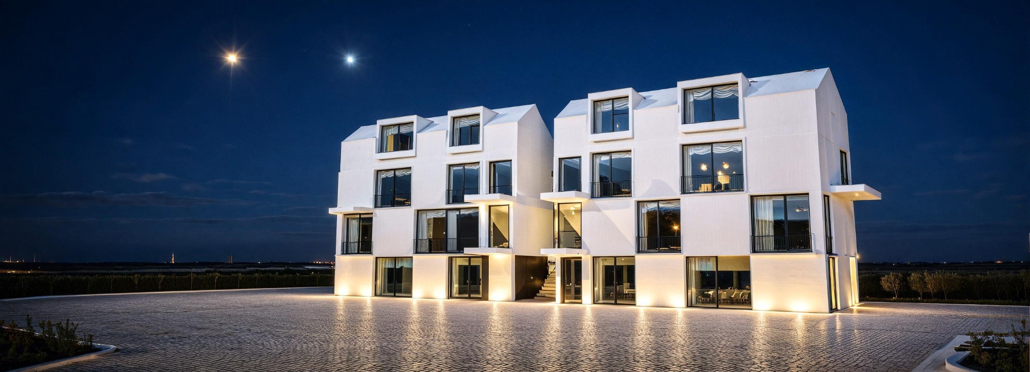 Nowoczesny budynek usługowy z białej elewacji oświetlony nocą, widoczne okna z balkonami i kamienna nawierzchnia przed budynkiem, w tle nocne niebo z gwiazdami.