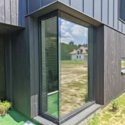 Nowoczesny dom z ciemną drewnianą elewacją i dużym narożnym oknem z żaluzjami poziomymi, odbijającym krajobraz z sąsiednim domem.
