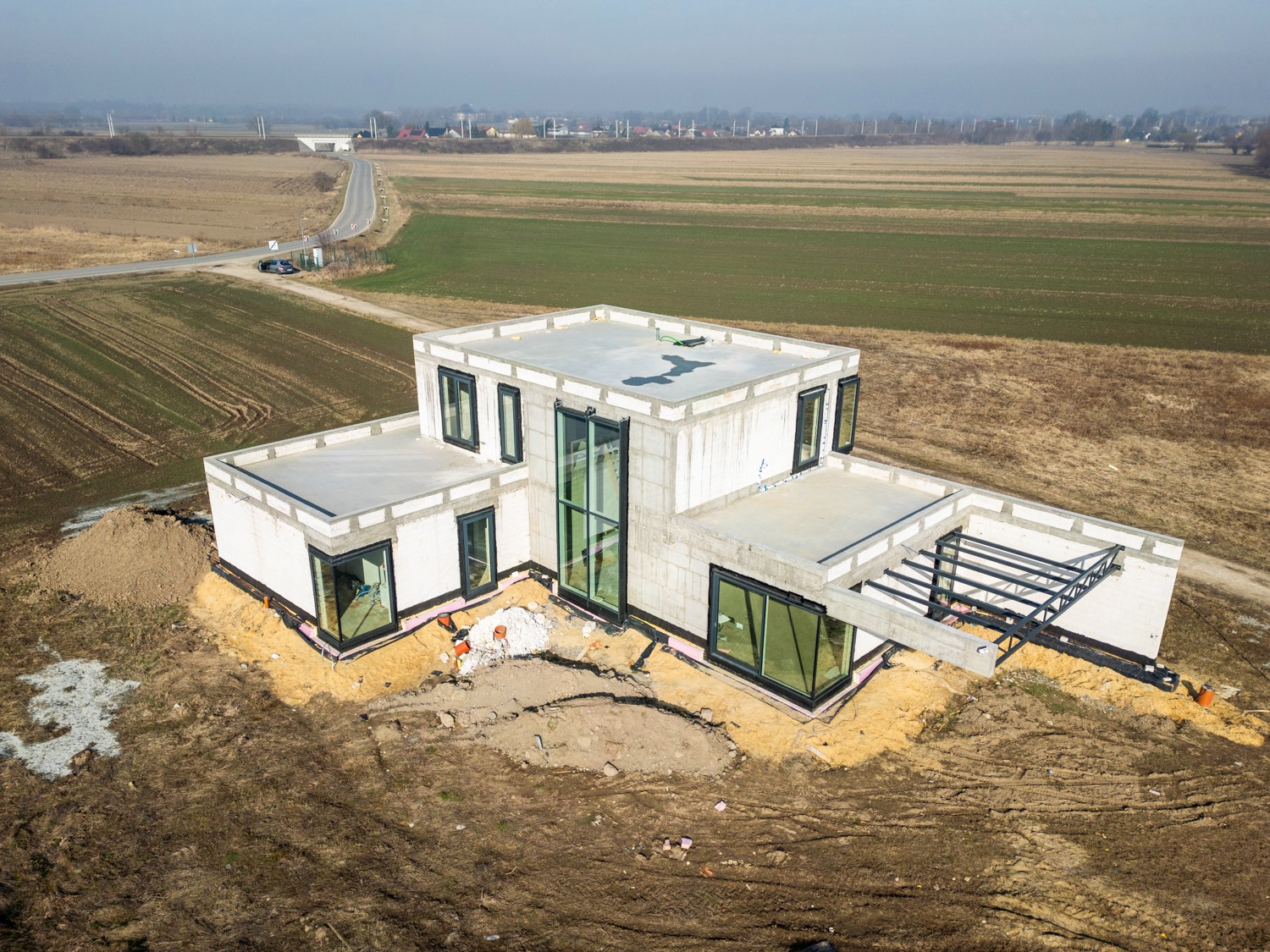 Surowy stan nowoczesnego domu z białego betonu komórkowego, z czarnymi oknami i przygotowaną konstrukcją zadaszenia, widok z drona na tle pól i drogi.