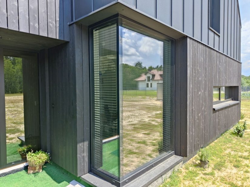 Nowoczesny dom z ciemną drewnianą elewacją i dużym narożnym oknem z żaluzjami poziomymi, odbijającym krajobraz z sąsiednim domem.