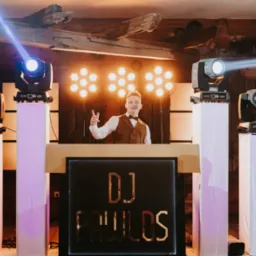 DJ w brązowej kamizelce i muszce, za podświetlonym stanowiskiem z napisem 'DJ PAWLOS', otoczony przez kolumny i ruchome głowy oświetleniowe na białych postumentach, z drewnianym sufitem w tle.