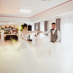 DJ Pawlos Łukasz Pawlos - Para młoda tańczy w dymie na tle sceny DJ-a z oświetleniem. Dekoracja sali weselnej z parkietem i światłami. Mężczyzna w muszce i kraciastej kamizelce, kobieta w białej sukni.