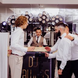 DJ Pawlos Łukasz Pawlos - DJ za konsoletą z efektownym oświetleniem, w otoczeniu elegancko ubranych gości, gotowych do zabawy. Ujęcie z imprezy, z bliska na interakcję z DJ-em.
