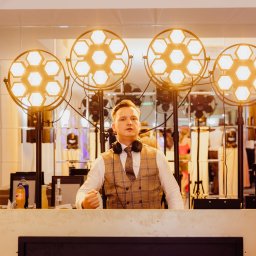 DJ Pawlos Łukasz Pawlos - DJ w kamizelce w kratę za konsoletą, otoczony przez cztery reflektory z ciepłym światłem i dwa reflektory z fioletowym światłem na tle lustrzanej ściany.