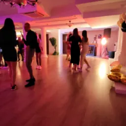 Tańczący ludzie na parkiecie podczas imprezy, w tle stanowisko DJ'a z podświetlanym panelem, dekoracje z balonów w kolorach czerni, bieli i złota.