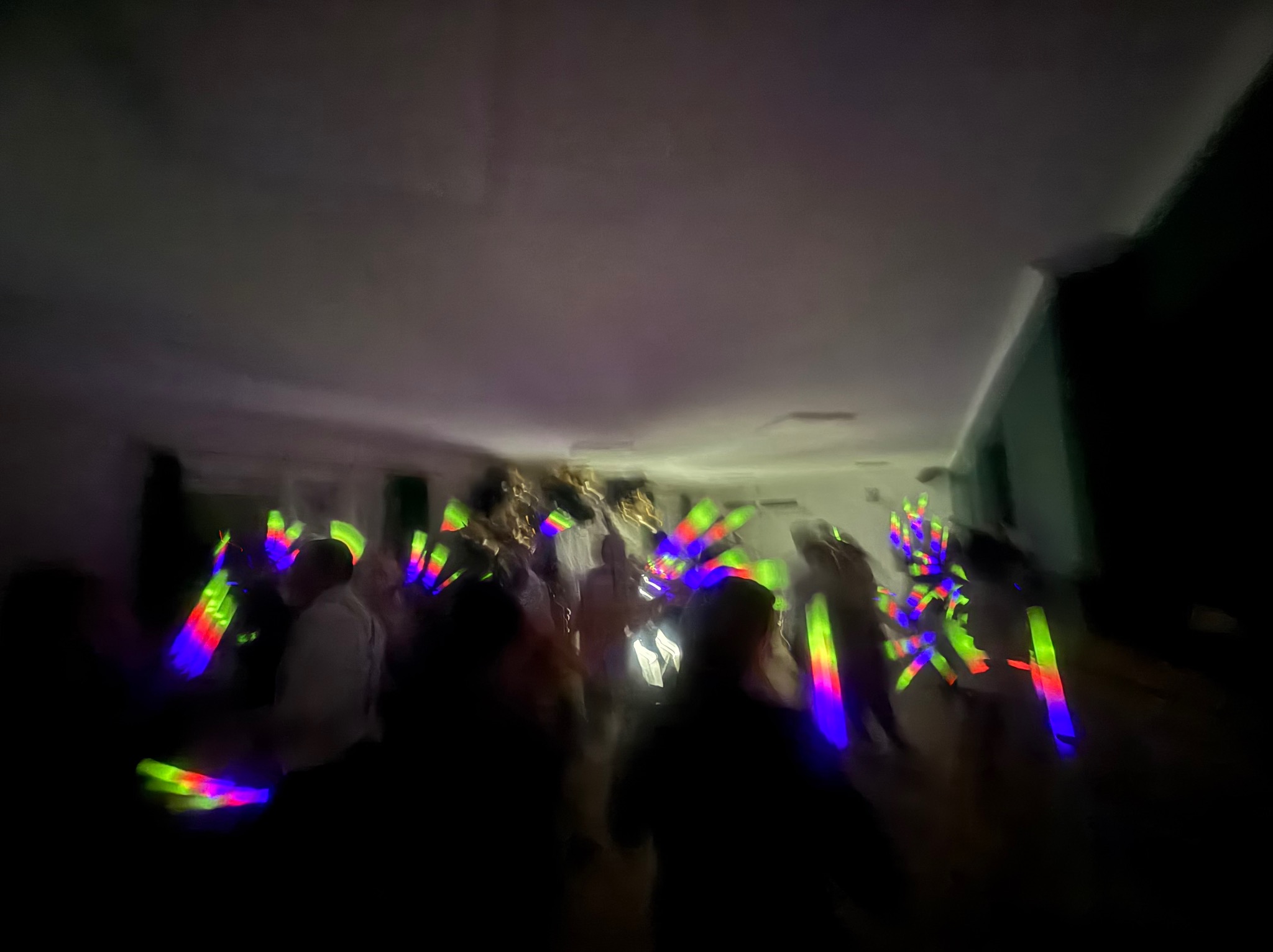 Rozmazane ujęcie tańczących osób z kolorowymi, świecącymi pałeczkami na imprezie w ciemnym pomieszczeniu. Dynamiczna atmosfera zabawy.