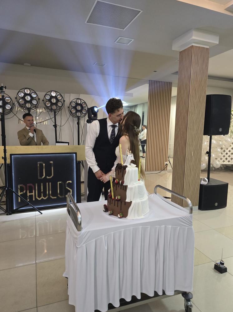 Para młoda całuje się przy torcie weselnym na tle sceny z DJ-em i nagłośnieniem. DJ w tle przy konsolecie, oświetlenie sceniczne, kolumna głośnikowa.