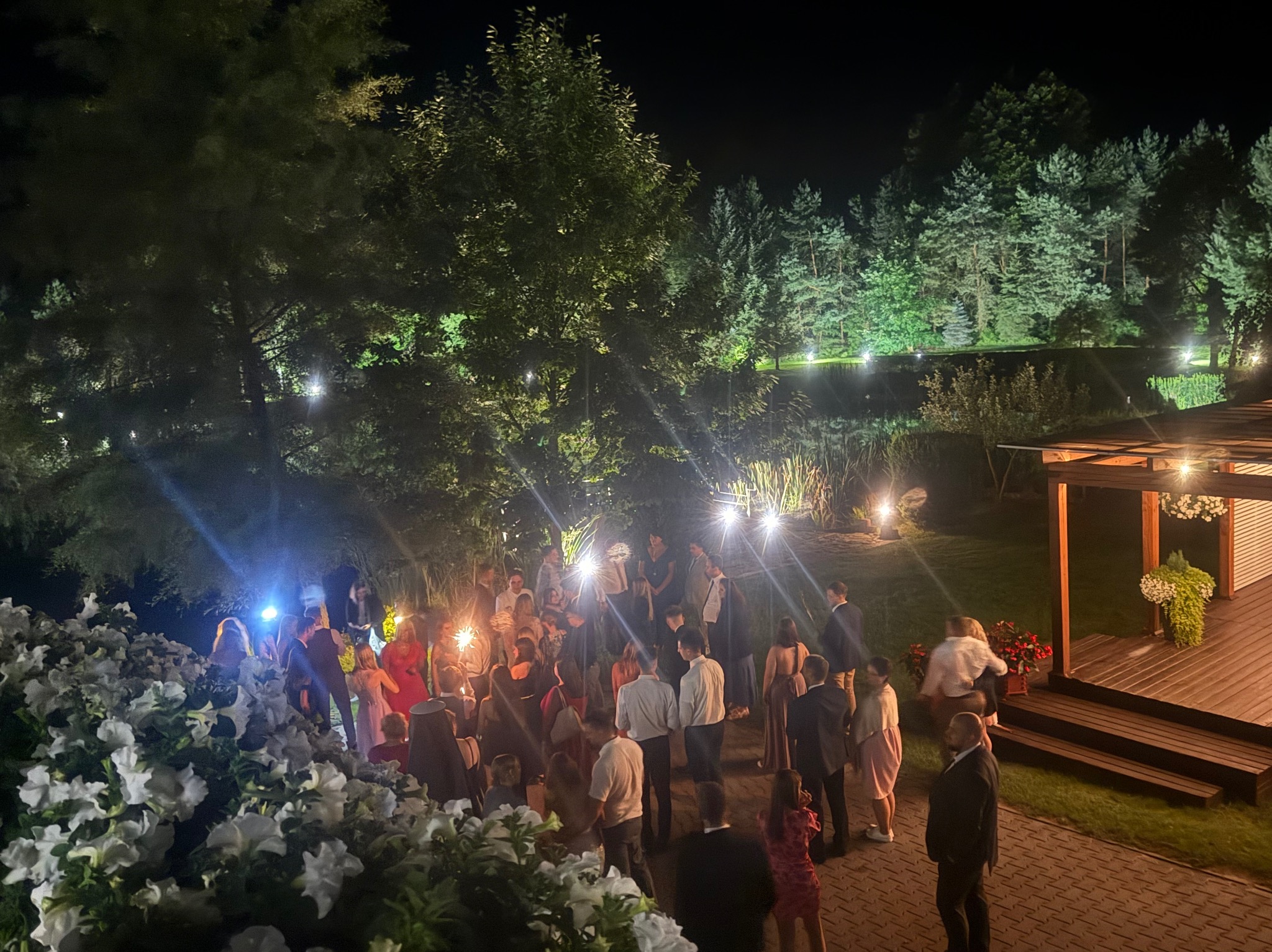 Nocna impreza plenerowa z gośćmi trzymającymi zimne ognie, oświetlona dekoracyjnymi lampkami, z widokiem na zalesiony teren i drewnianą altanę.