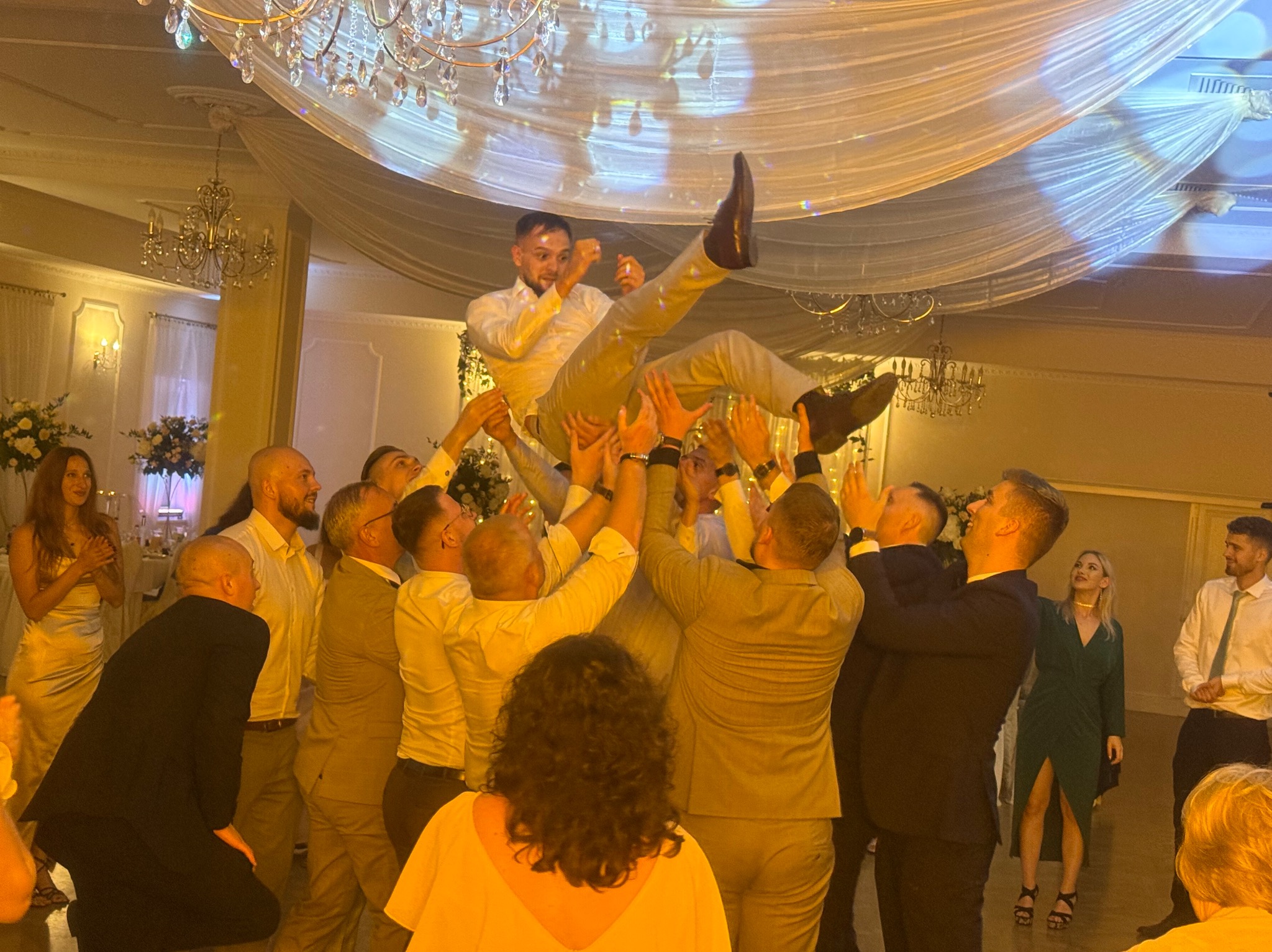 Radosna scena weselna: grupa mężczyzn podrzuca pana młodego w górę, w tle widoczni goście i dekoracje sali balowej z żyrandolami.