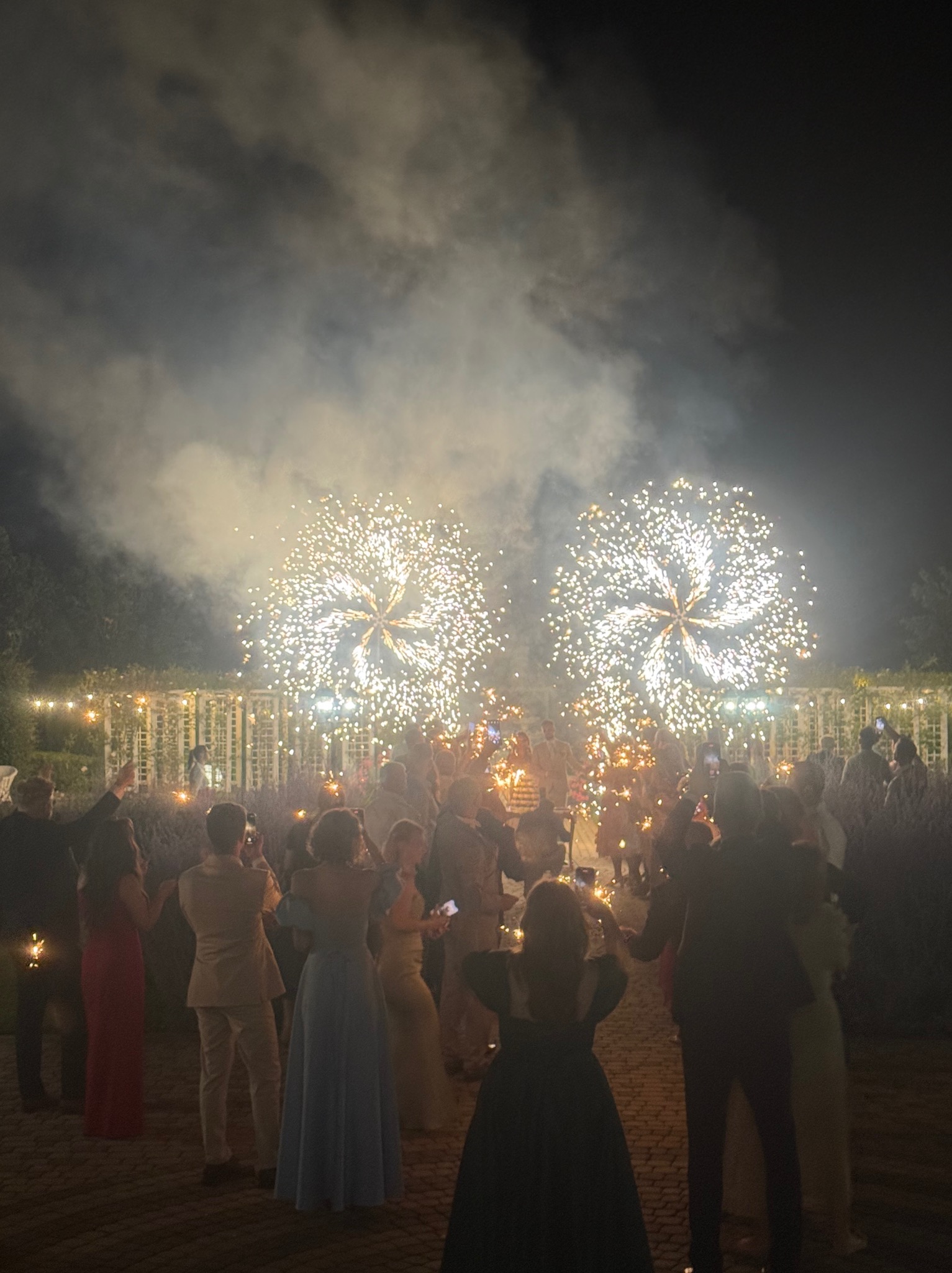 Nocne świętowanie z fajerwerkami i iskrzącymi się zimnymi ogniami w rękach gości, uchwycone z dołu podczas imprezy plenerowej.