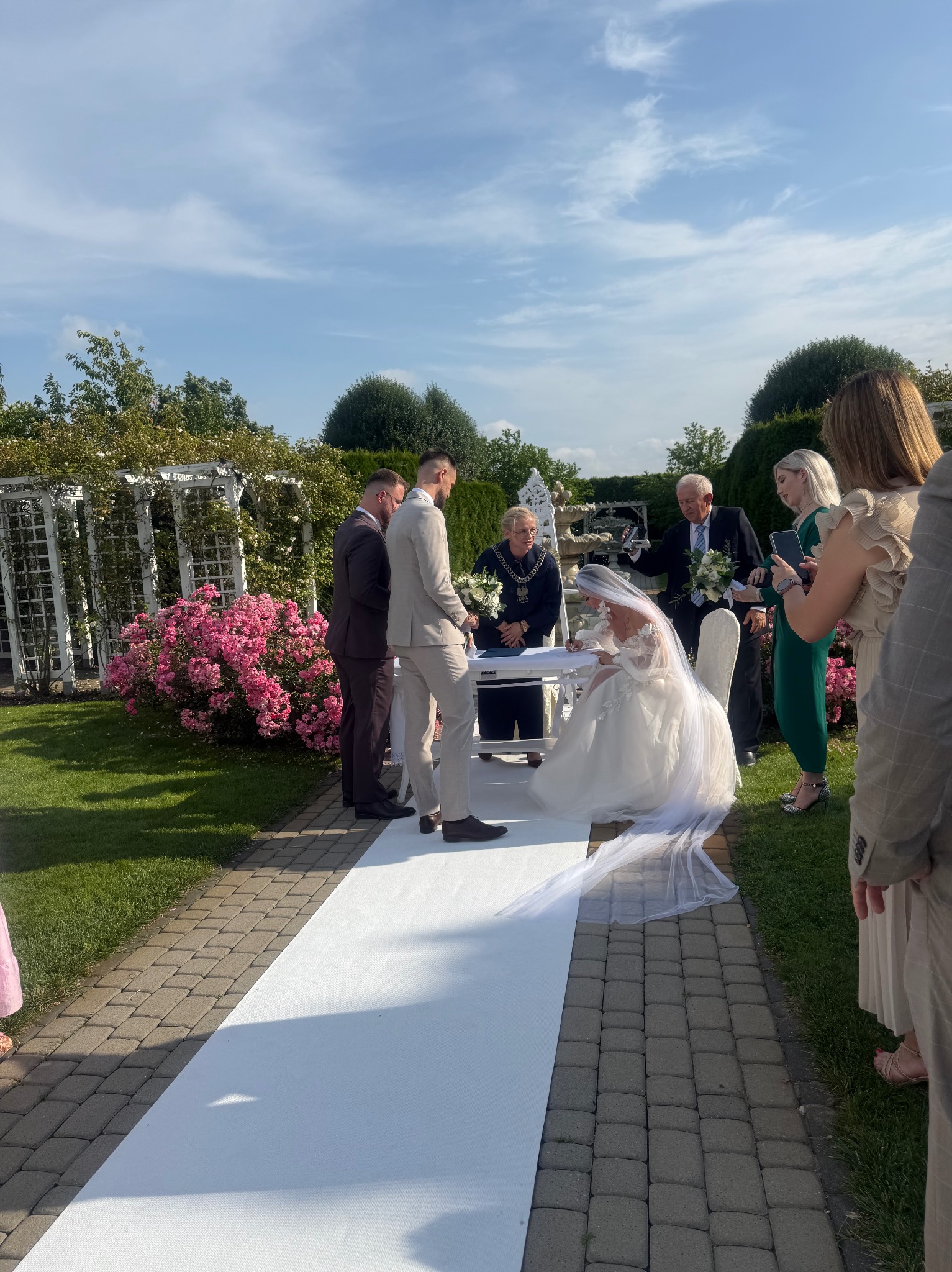 Ceremonia ślubna w plenerze: panna młoda klęczy podpisując dokument, otoczona urzędnikiem, świadkami i gośćmi na tle zielonego ogrodu z białym dywanem.