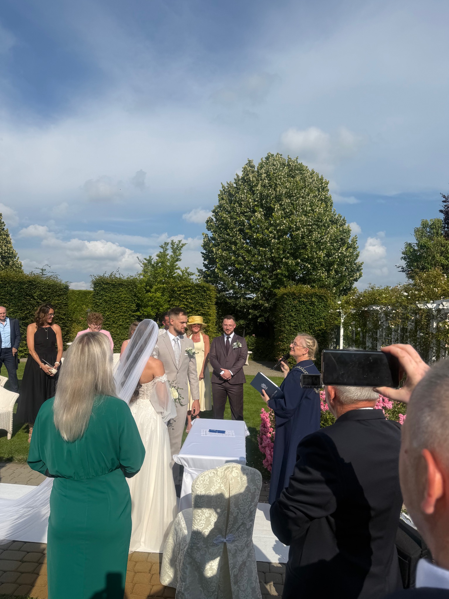 Plenerowa ceremonia ślubna: para młoda przed urzędnikiem, goście w tle, osoba filmująca smartfonem. Uroczystość w ogrodzie, słoneczny dzień.
