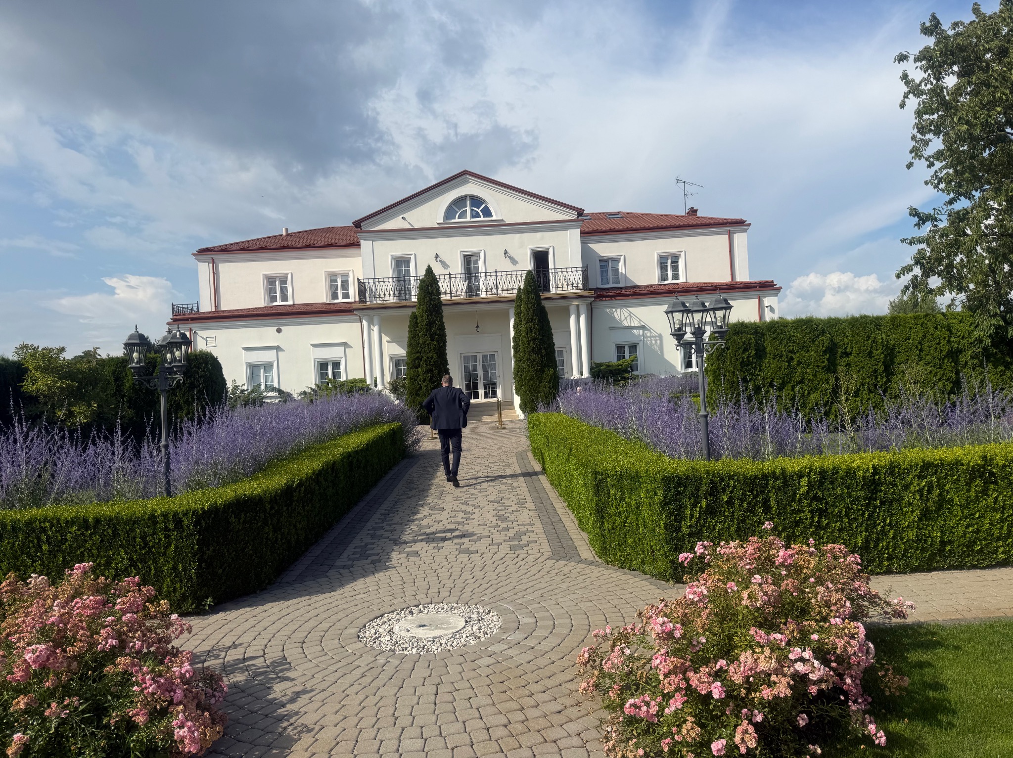 Elegancki dwór z czerwonym dachem, zadbany ogród z równo przyciętymi żywopłotami i kwitnącymi rabatami. Mężczyzna w garniturze idzie w stronę wejścia.