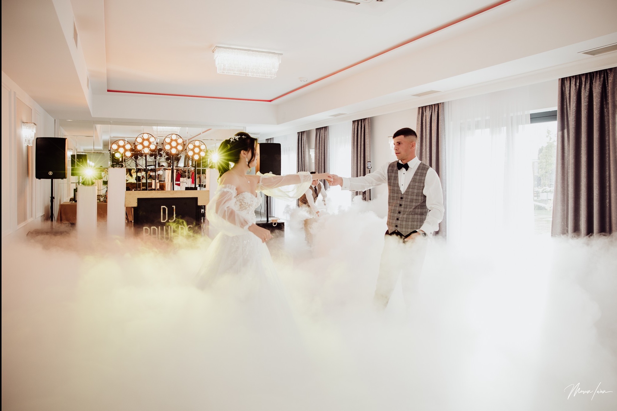 Para młoda tańczy w dymie na tle sceny DJ-a z oświetleniem. Dekoracja sali weselnej z parkietem i światłami. Mężczyzna w muszce i kraciastej kamizelce, kobieta w białej sukni.