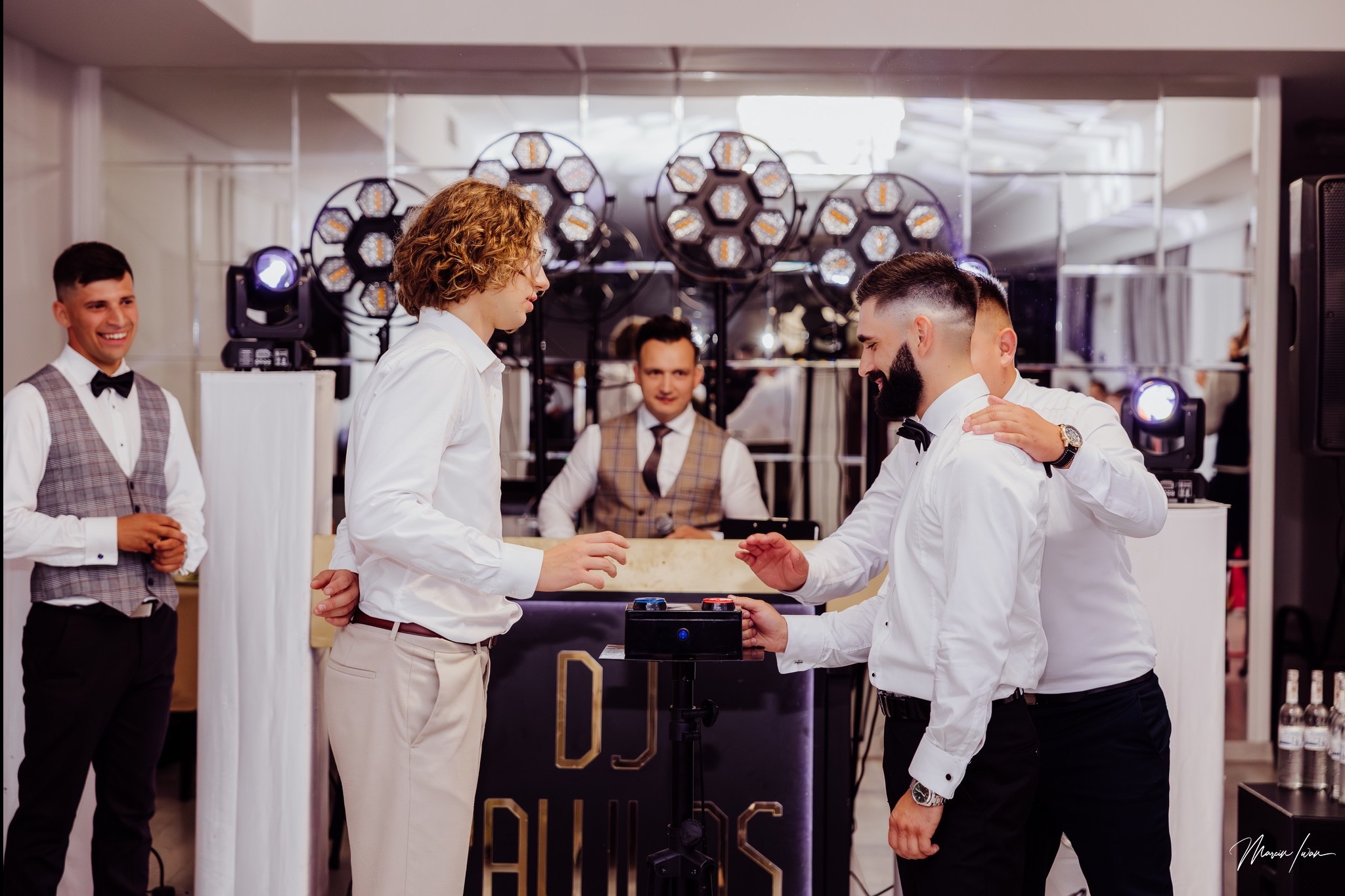 DJ za konsoletą z efektownym oświetleniem, w otoczeniu elegancko ubranych gości, gotowych do zabawy. Ujęcie z imprezy, z bliska na interakcję z DJ-em.