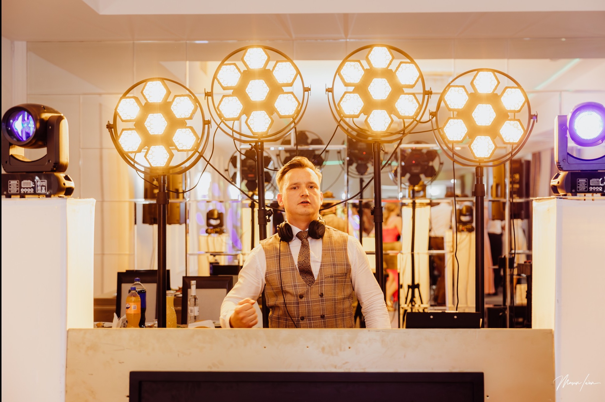 DJ w kamizelce w kratę za konsoletą, otoczony przez cztery reflektory z ciepłym światłem i dwa reflektory z fioletowym światłem na tle lustrzanej ściany.