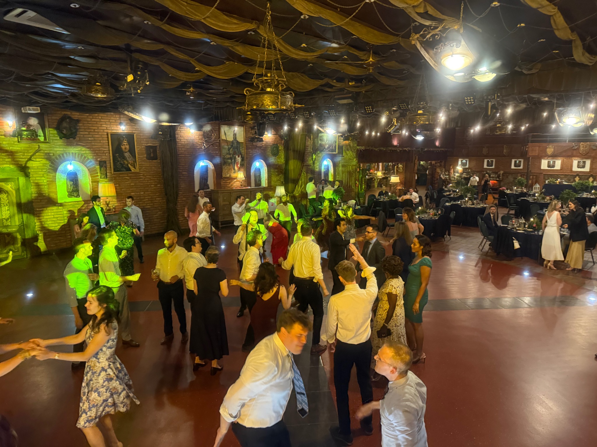 Sala balowa w stylu zamkowym z tańczącymi gośćmi, oświetlona zielonymi reflektorami i zdobiona żyrandolami. Ustawione stoły z nakryciami w tle.