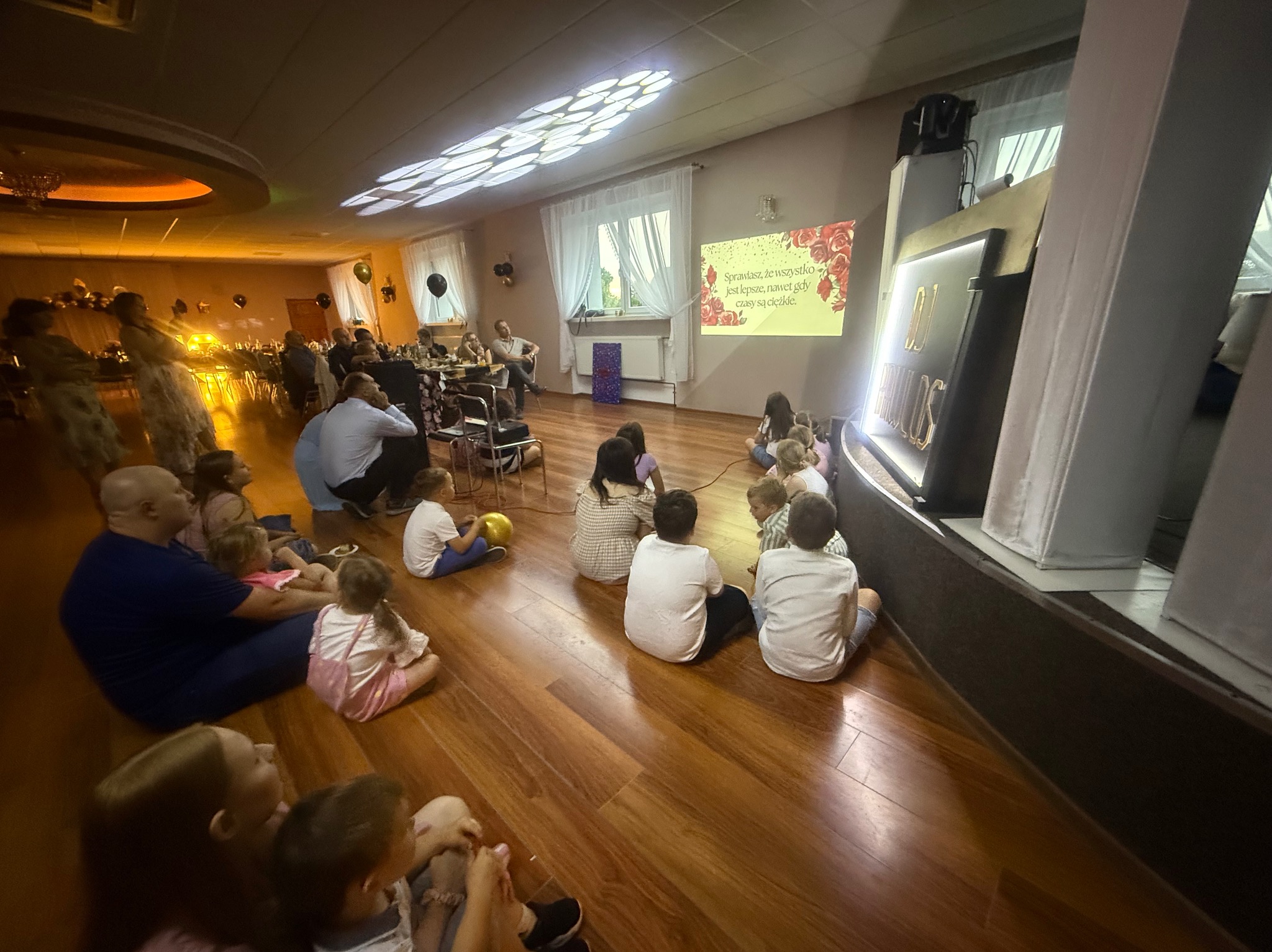 Sala bankietowa z dziećmi siedzącymi na podłodze, oglądającymi prezentację wyświetlaną na ekranie. W tle widoczny stół z gośćmi i dekoracje.