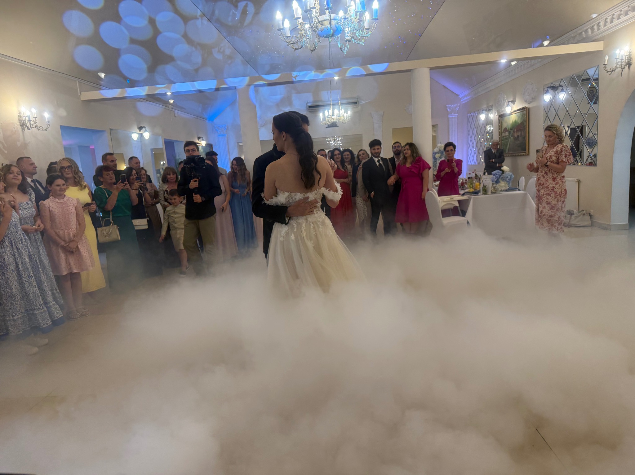 Para młoda w tańcu w chmurach dymu na sali weselnej, goście obserwują. Dekoracje świetlne na suficie, kolumny i lustra w tle. Romantyczna atmosfera.