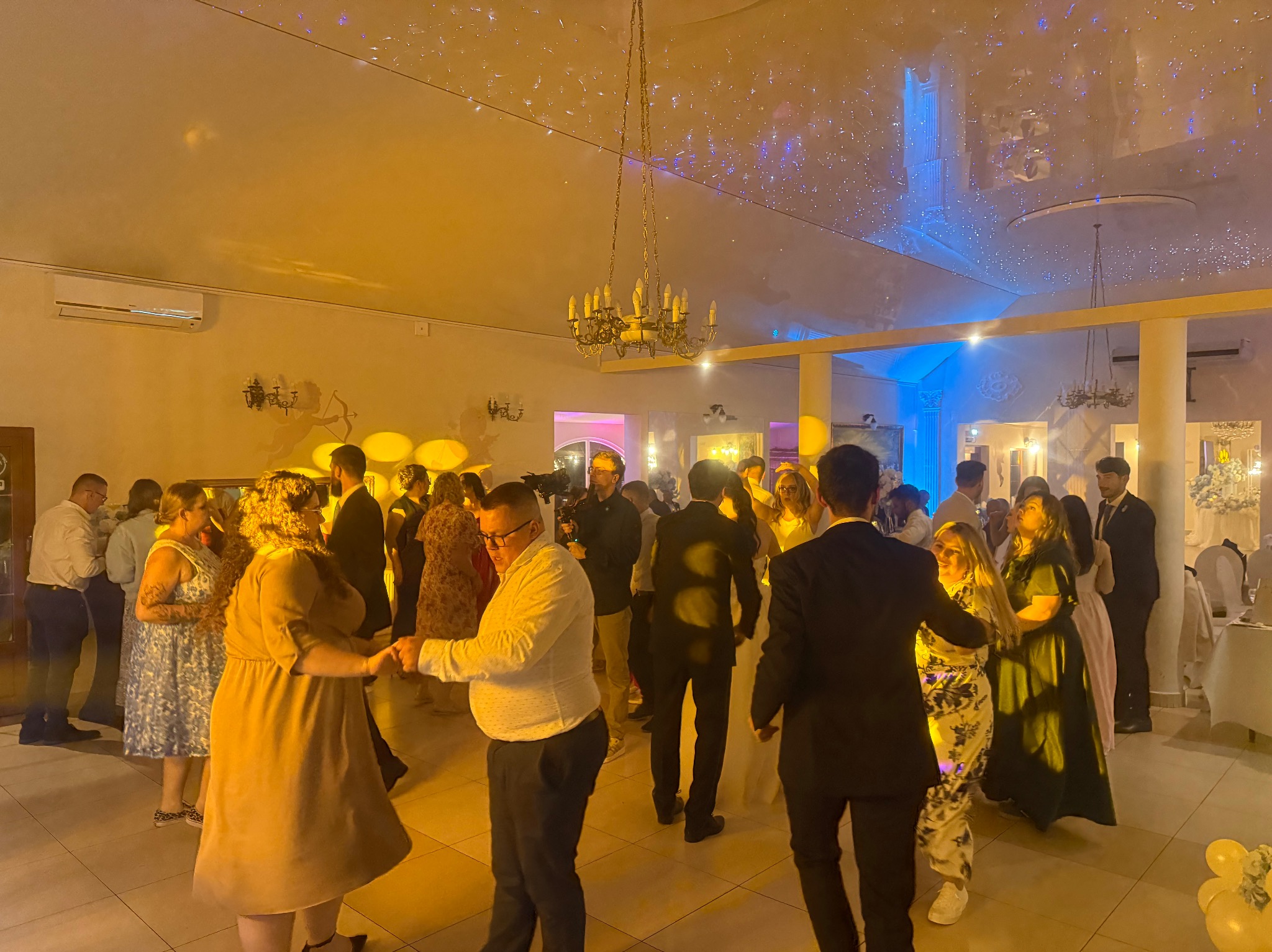 Tańczący goście weselni w sali z dekoracyjnym oświetleniem sufitowym i żyrandolem. Widoczni ludzie w różnym wieku, w strojach wieczorowych, w ruchu podczas zabawy.