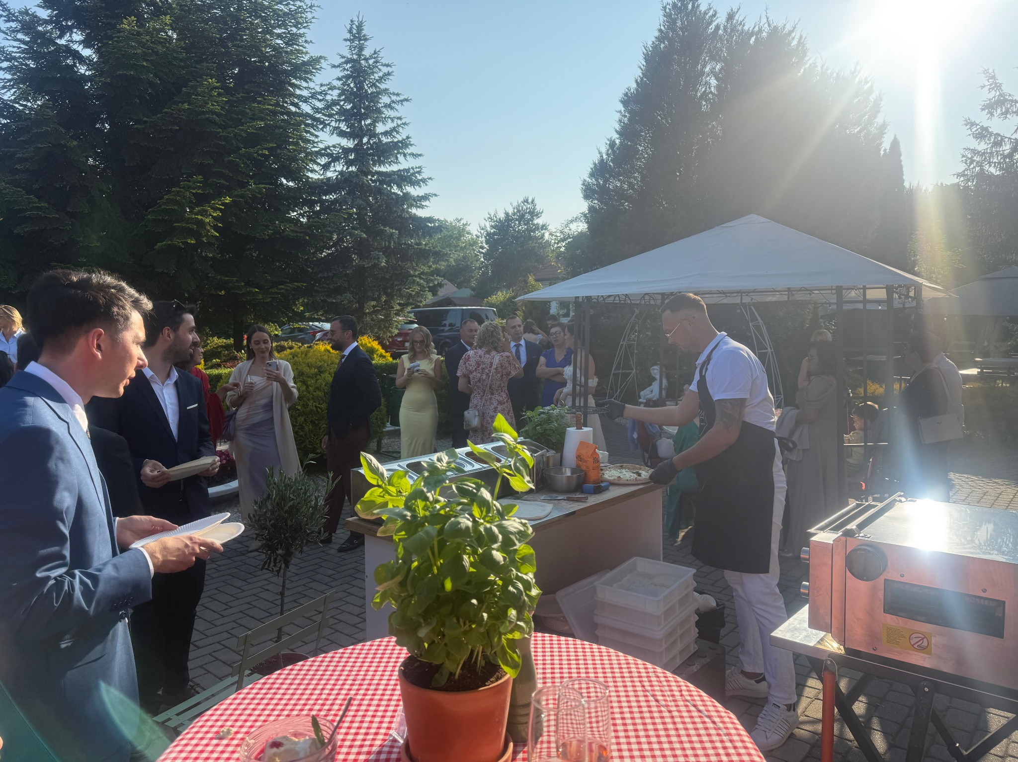 Plenerowa stacja do pizzy na przyjęciu: kucharz w fartuchu przygotowuje pizzę dla gości. W tle ogród i biały namiot.