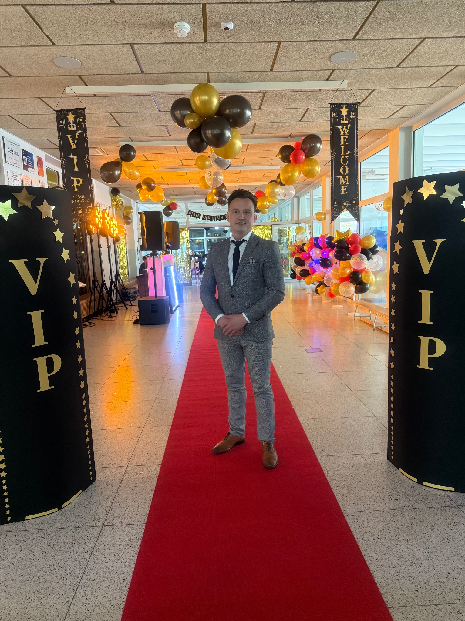 Mężczyzna w garniturze stoi na czerwonym dywanie, otoczony dekoracjami VIP z balonami i oświetleniem, sugerującymi wejście na imprezę lub wydarzenie.