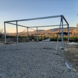 Metalowa konstrukcja wiaty na żwirowym podłożu, w tle krajobraz z pagórkami i drzewami o jesiennych liściach, widok w trakcie budowy.