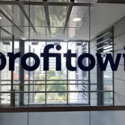 Niebieski napis 'profitowi' na szklanej ścianie biurowca z widokiem na miasto w tle.