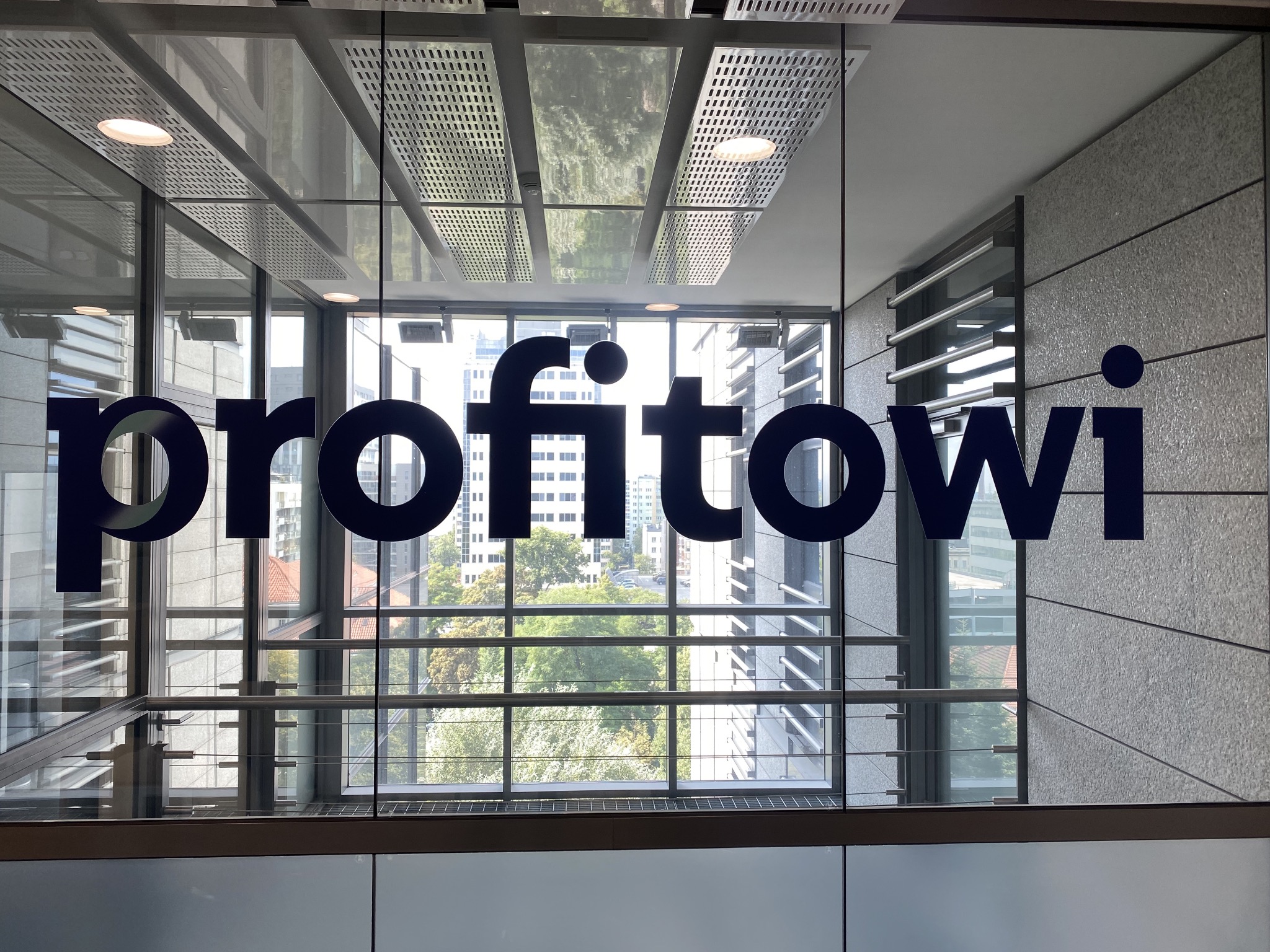 Niebieski napis 'profitowi' na szklanej ścianie biurowca z widokiem na miasto w tle.