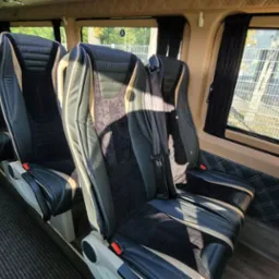 Wnętrze luksusowego busa z rzędem czarno-beżowych skórzanych foteli z welurowymi wstawkami, widok z perspektywy pasażera na okno z zasłonką i napis 'Wyjście awaryjne'.