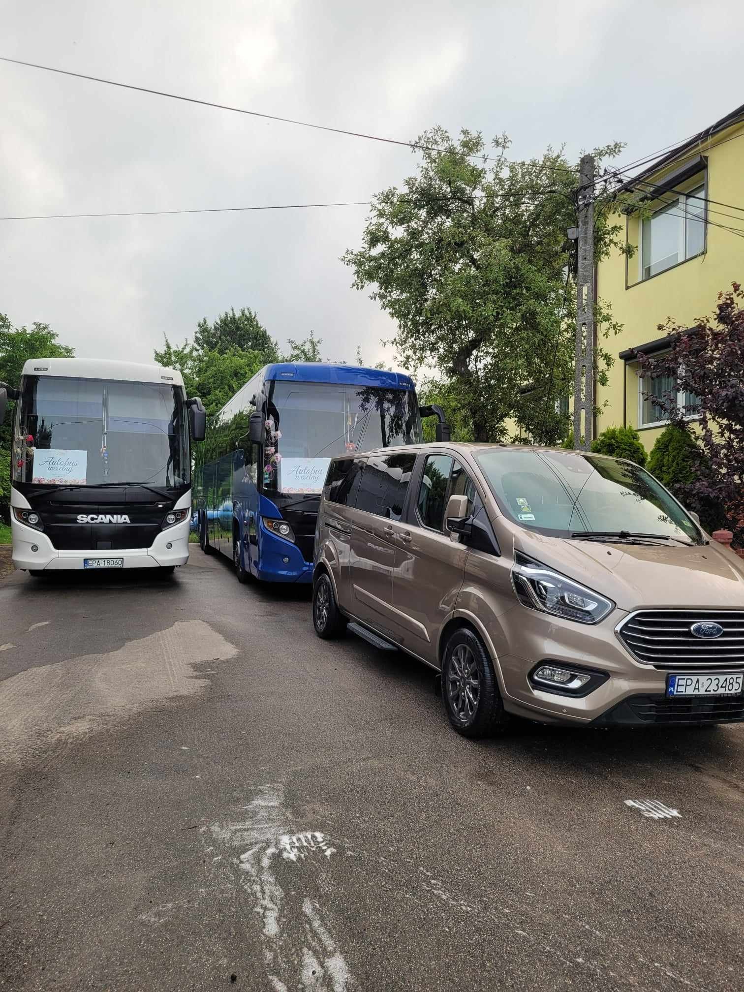 Dwa autokary Scania z dekoracjami ślubnymi i brązowy van Ford Transit zaparkowane na mokrej ulicy przed żółtym budynkiem w pochmurny dzień.