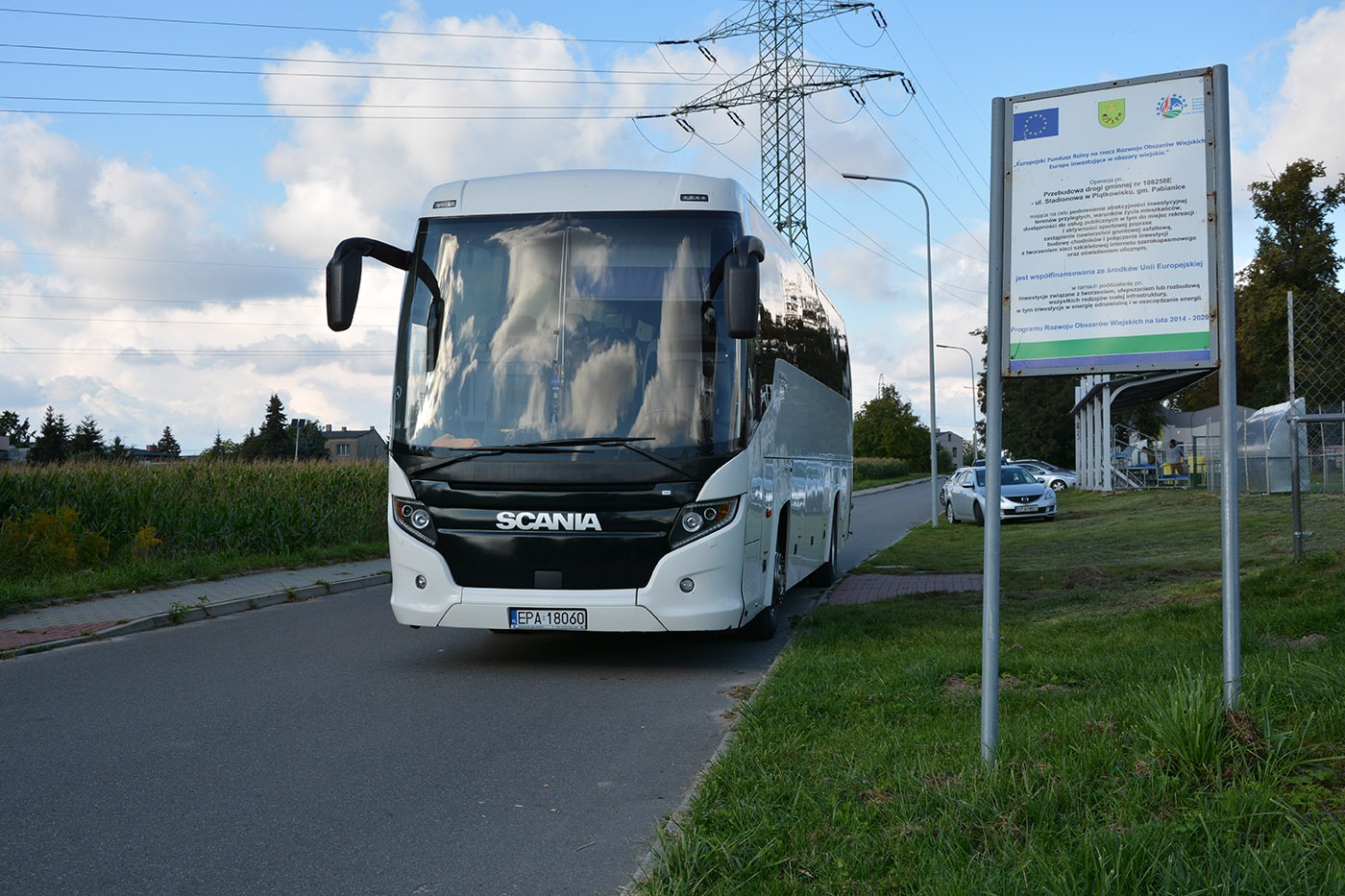Biały autokar marki Scania zaparkowany na poboczu drogi, z odbiciem chmur na przedniej szybie, w tle słup wysokiego napięcia i tablica informacyjna o funduszach europejskich.