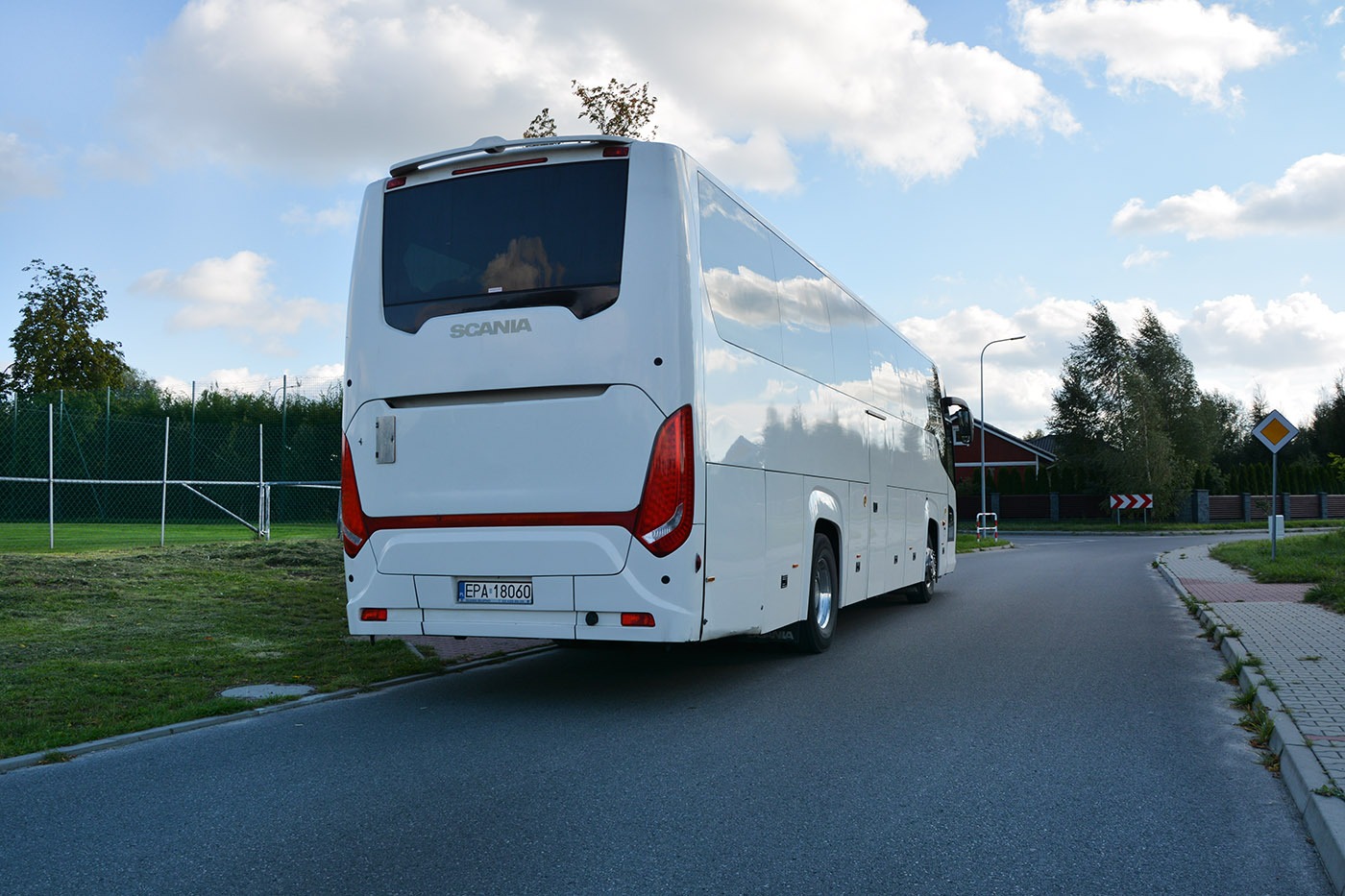 Biały autokar marki Scania, widziany z tyłu, zaparkowany na asfaltowej drodze w słoneczny dzień. Widoczne tablice rejestracyjne oraz elementy oświetlenia.