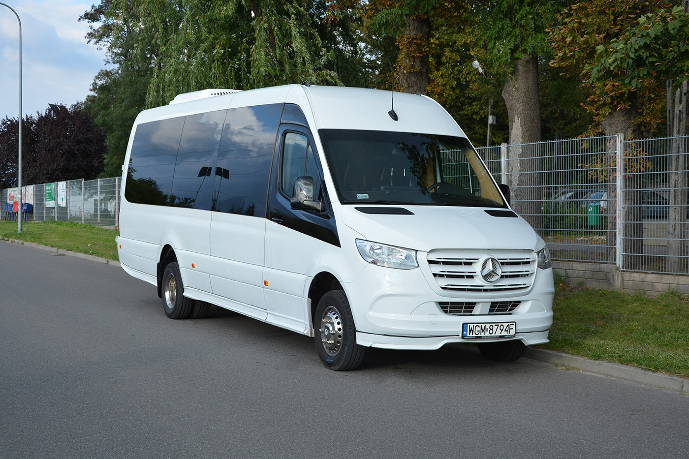 Biały Mercedes-Benz Sprinter z przyciemnianymi szybami, zaparkowany na asfaltowej drodze przy chodniku i metalowym ogrodzeniu.