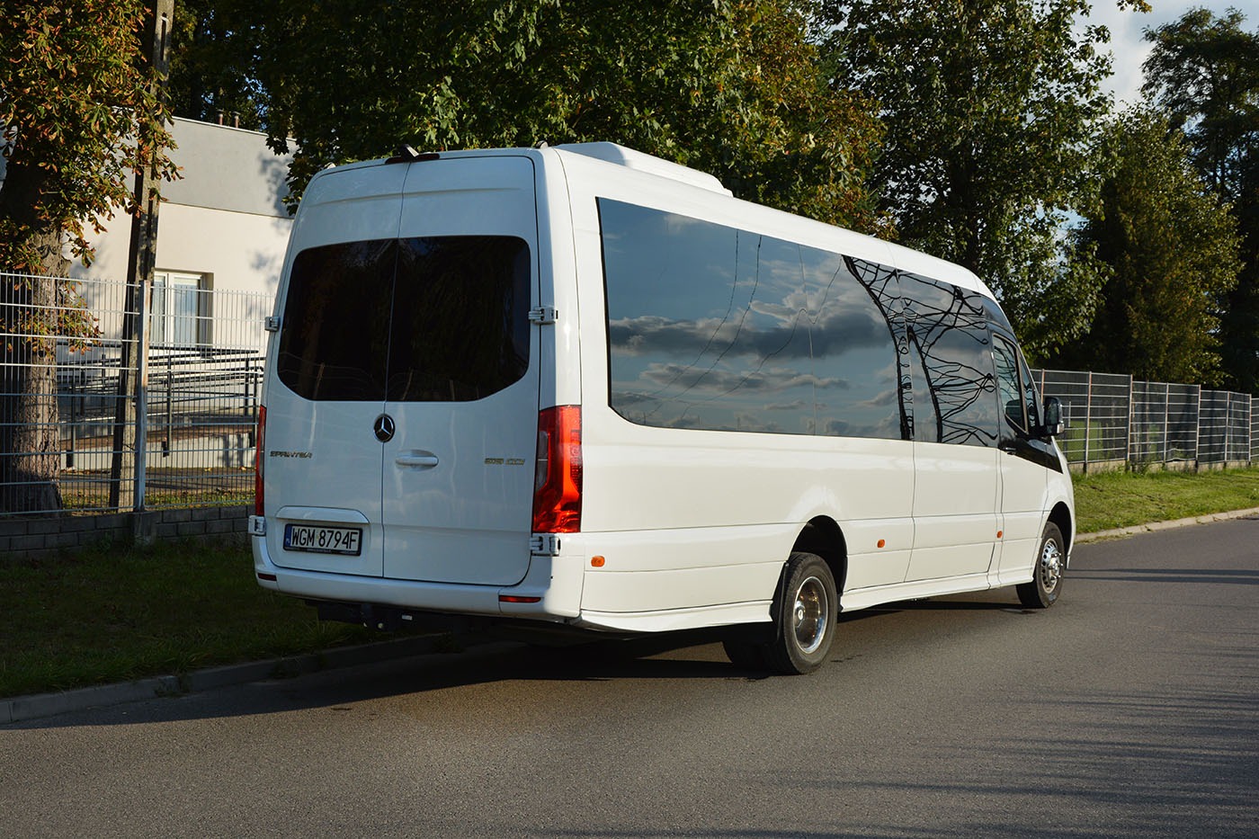 Biały minibus Mercedes-Benz Sprinter z przyciemnianymi szybami, zaparkowany na poboczu drogi, z widocznym odbiciem chmur w oknach.