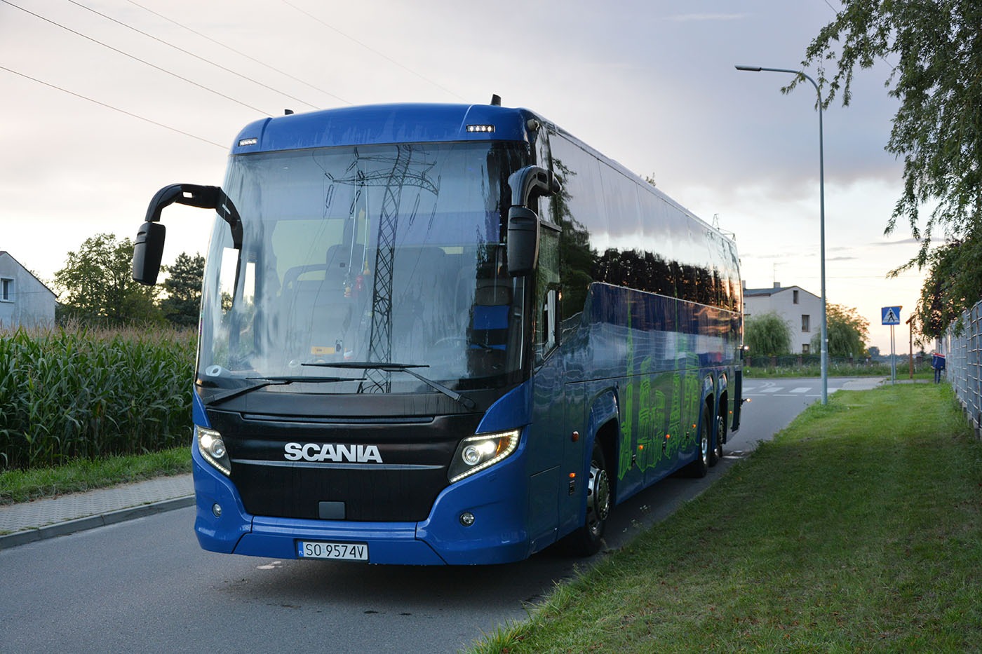 Niebieski autokar marki Scania zaparkowany na poboczu drogi, z widocznym odbiciem słupów wysokiego napięcia w przedniej szybie.