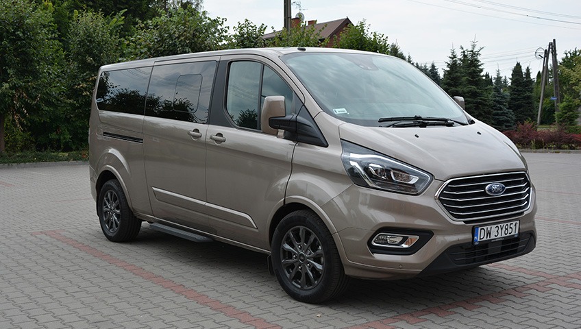 Beżowy Ford Transit Custom na parkingu z kostki brukowej, widok z przodu i boku, z widoczną tablicą rejestracyjną i logo Forda.
