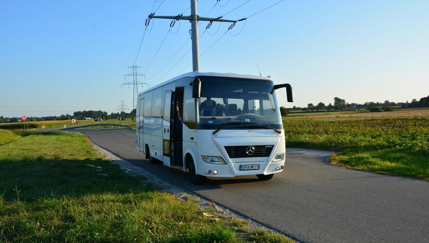 Biały minibus Mercedes-Benz z otwartymi drzwiami stoi na asfaltowej drodze na tle pól i linii energetycznych, w słoneczny dzień.