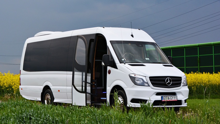 Biały, luksusowy Mercedes Sprinter z otwartymi drzwiami, zaparkowany na zielonej trawie z kwitnącym polem rzepaku w tle.