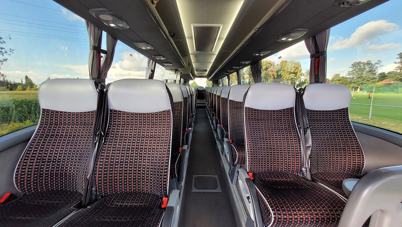 Wnętrze autobusu turystycznego z rzędami foteli z brązowo-czarną tapicerką w geometryczny wzór, widok na zewnątrz przez okna z zasuniętymi zasłonami, na zewnątrz widoczny krajobraz z zielenią...