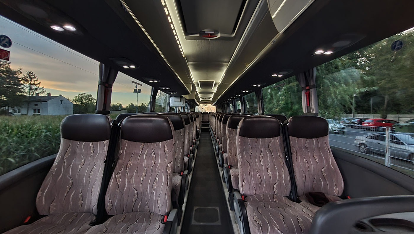 Wnętrze autobusu z rzędami foteli obitych wzorzystą tapicerką, widok z perspektywy kierowcy, za oknem krajobraz z budynkiem i zaparkowanymi samochodami.