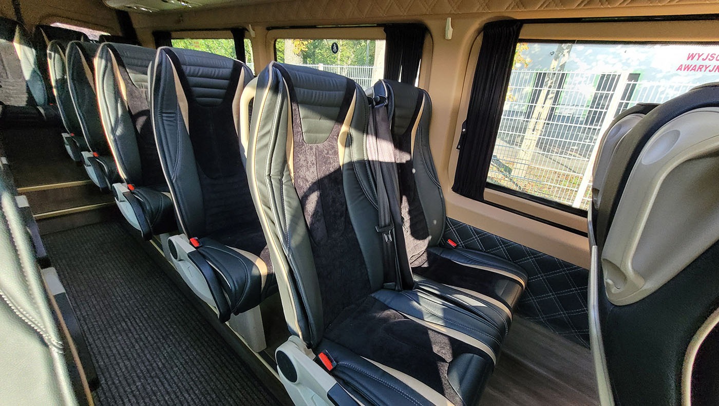 Wnętrze luksusowego busa z rzędem czarno-beżowych skórzanych foteli z welurowymi wstawkami, widok z perspektywy pasażera na okno z zasłonką i napis 'Wyjście awaryjne'.