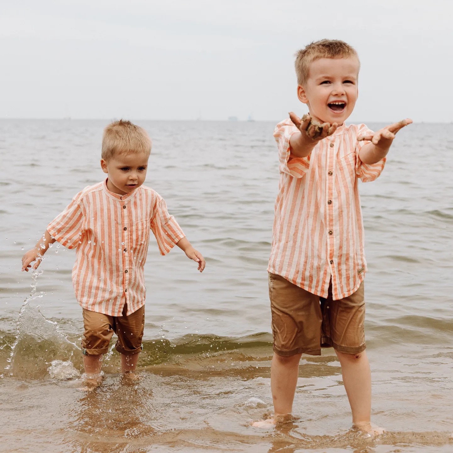 Dwóch chłopców w identycznych koszulach w paski i brązowych spodenkach bawi się w płytkiej wodzie morskiej; jeden z chłopców trzyma w dłoniach mokry piasek, uśmiechając się szeroko, drugi wchodzi...