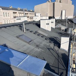 Valentyn Verbitskyi Dekarz - Czarny dach pokryty papą termozgrzewalną z lukarnami i kominami wentylacyjnymi na tle jasnych budynków w słoneczny dzień. Widoczne elementy rusztowania.