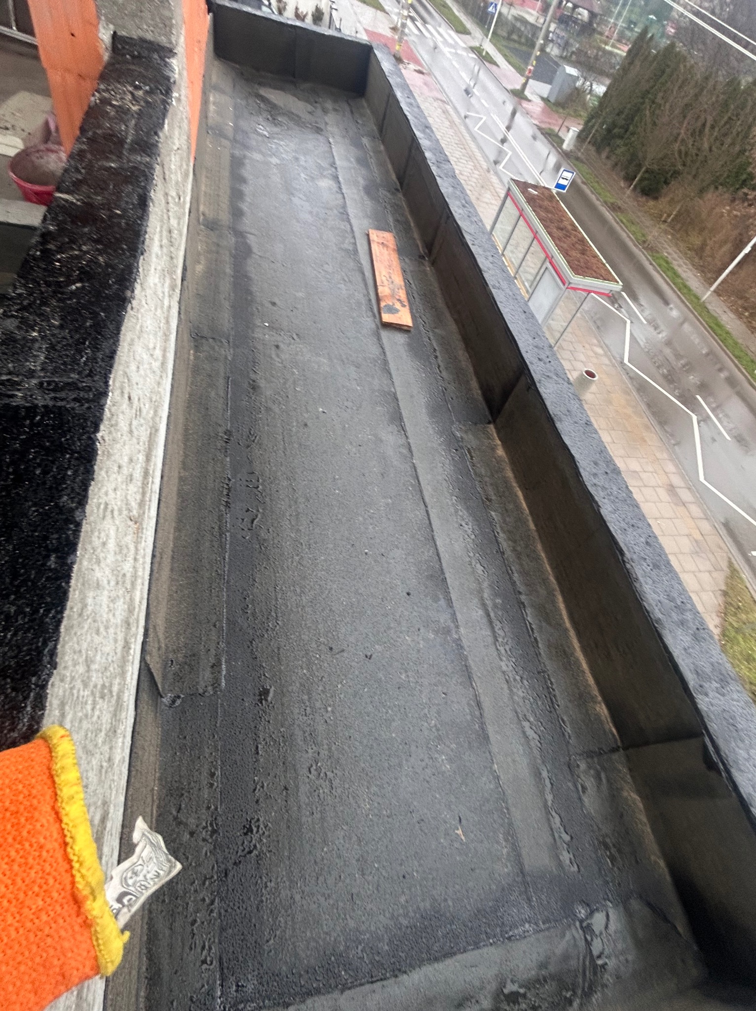 Uszczelniony, płaski dach z papy bitumicznej, widok z góry. W tle fragment ulicy i przystanek autobusowy. Pomarańczowa rękawica robocza w lewym dolnym rogu kadru.